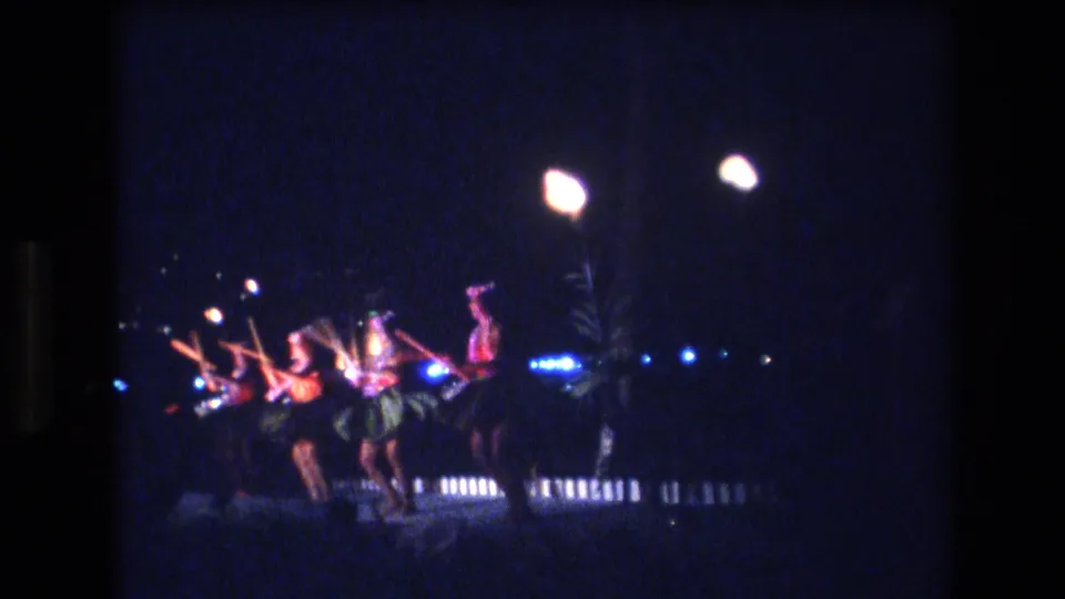 1977 Honolulu, Oahu Super 8 frame showing a synchronized Hawaiian dance routine in low light with palm fronds and a fence behind the performers.
