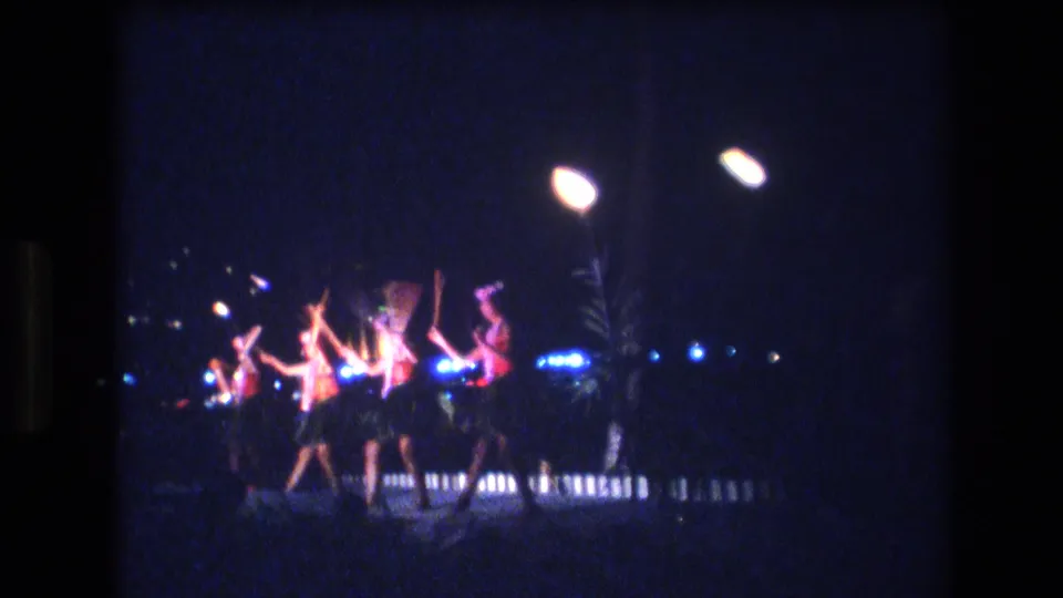 1977 Honolulu, Oahu home-movie frame of women in a nighttime Hawaiian line dance, with strong stage lighting and visible film grain.