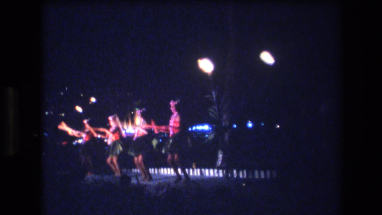 In 1977 Honolulu, four women in red and dark performance costumes appear to dance in formation on a night stage with bright lamps and dark foliage behind them.