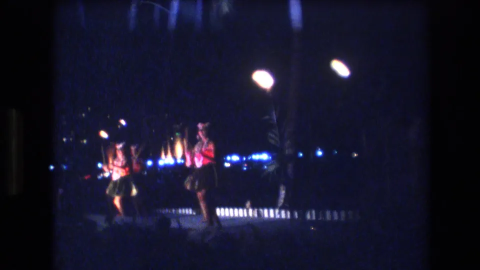 The 8mm frame shows the dance group shifting position on the night stage, with red costume details, bright lamps, and soft low-light film grain.