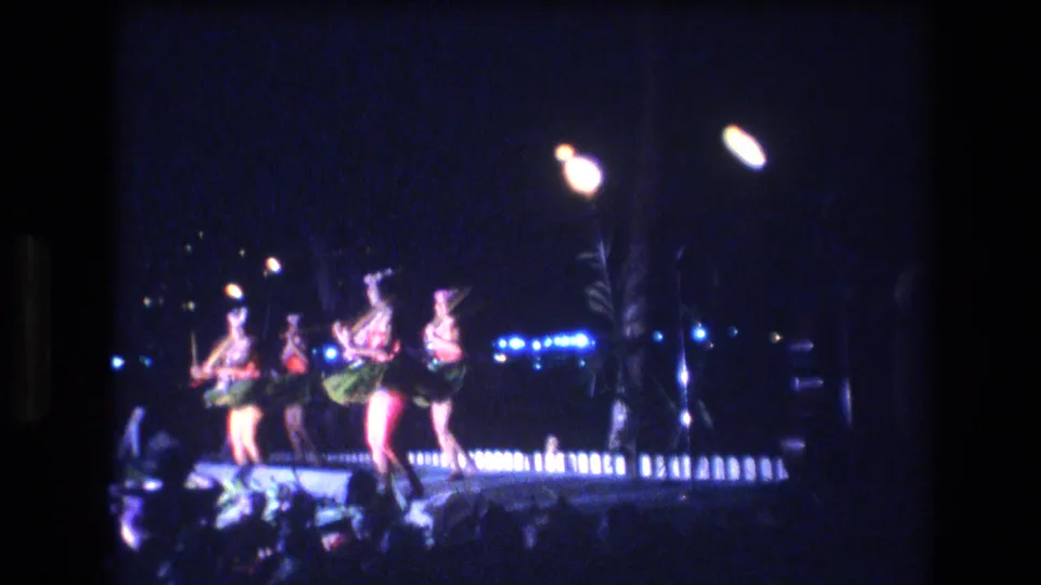 Four women in red tops, dark bottoms, green skirt elements, and headpieces dance in a line on a Honolulu night stage, framed by bright lamps and a dark tropical backdrop.