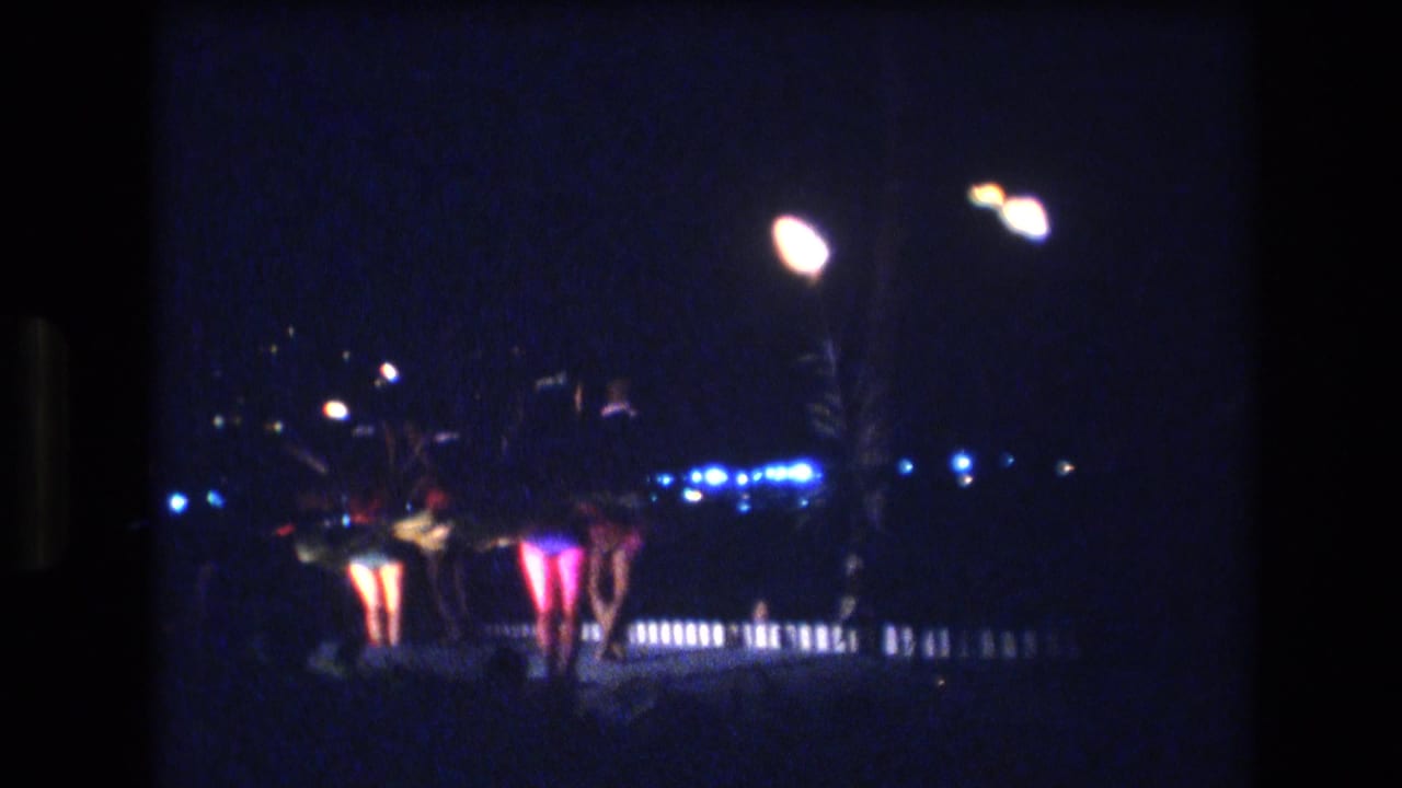 1977 Honolulu home-movie frame showing hula dancers on a nighttime stage, seen from the audience under bright white lamps and blue background lighting.