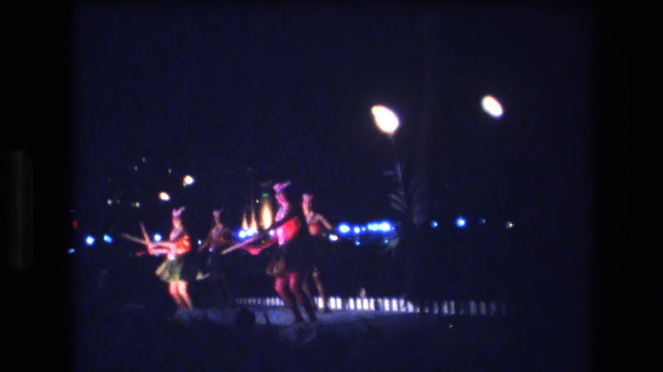1977 Honolulu frame of hula dancers in grass skirts and bright costume colors performing beneath overhead lamps and blue stage lights at night.