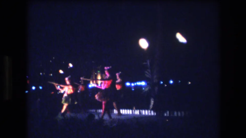 1977 Honolulu home-movie frame showing dancers in grass skirts moving across a low-lit outdoor stage with bright lamps, blue lights, and audience silhouettes nearby.