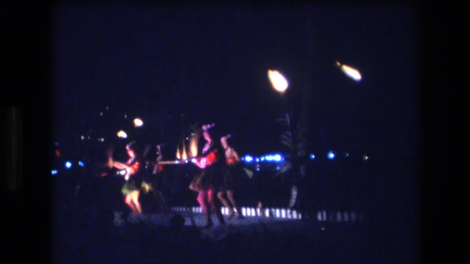 1977 Honolulu frame of hula dancers grouped on a nighttime stage, with bright overhead lamps, blue background lights, and soft 8mm grain visible throughout.