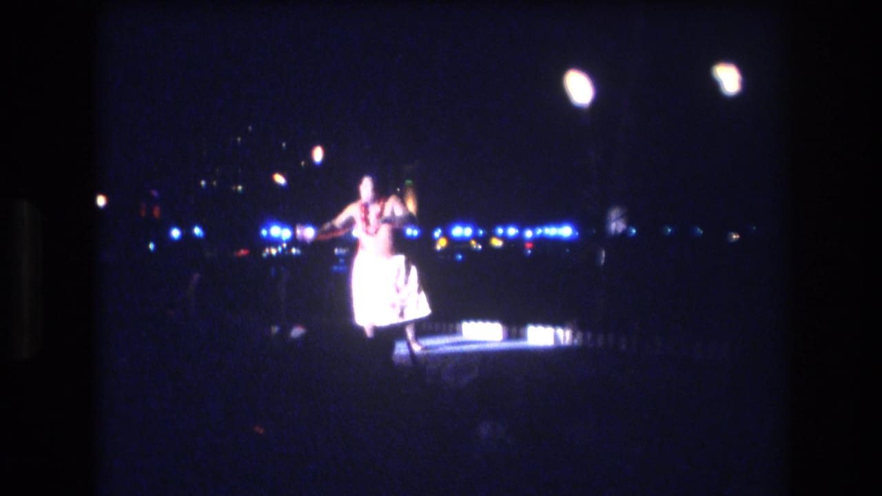 In metadata-supported 1977 Honolulu, a hula dancer performs on a dim outdoor stage, surrounded by blue-white lights and torch-like flames in soft 8mm focus.