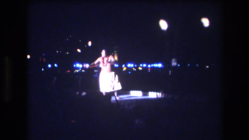 In metadata-supported 1977 Honolulu, a hula dancer performs on a dim outdoor stage, surrounded by blue-white lights and torch-like flames in soft 8mm focus.