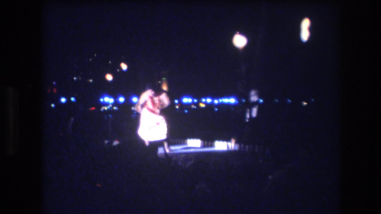 Hula dancer bends forward on a dim stage with rows of lights in the background.