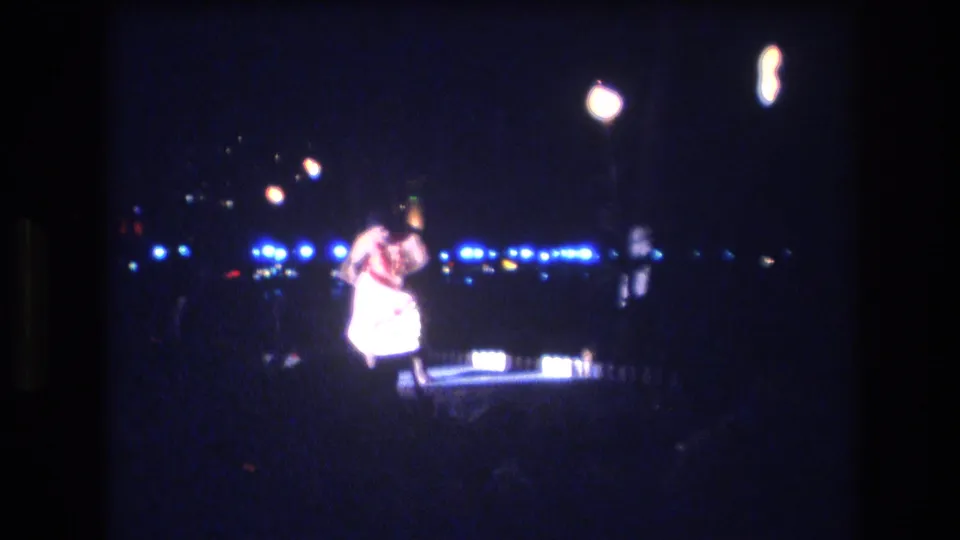 The dancer continues the outdoor hula performance in Honolulu, leaning into the movement as small stage lights and torch flames glow across the dark background.
