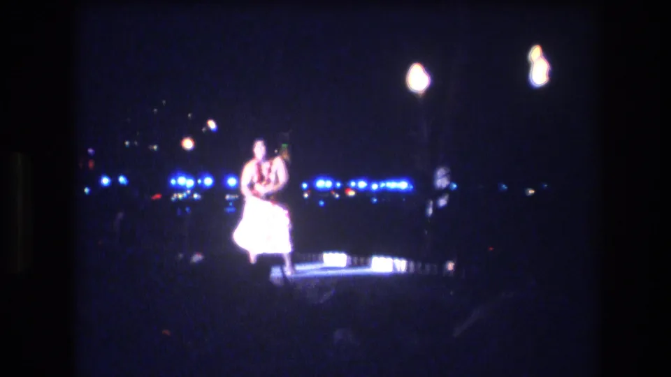 A centered view shows the hula dancer on the night stage in Honolulu, with her light costume catching the exposure while background lights and flames remain softly blurred.