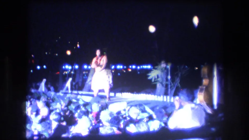 A wider 1977 Honolulu home-movie frame shows the hula dancer on a decorated outdoor stage, with bright edge lighting, tropical foliage, torch-like flames, and a possible guitar or prop at right.