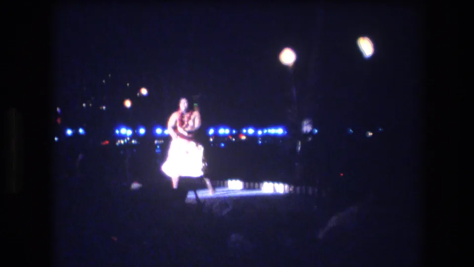 A male hula dancer in white attire and a lei performs on a small nighttime stage in Honolulu, Oahu, Hawaii, as supported by the clip metadata.
