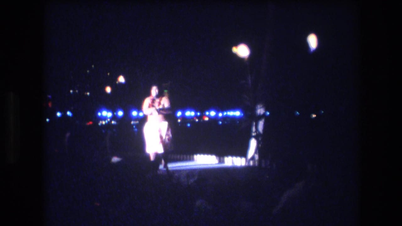 Male performer stands centered on a dark stage with bright lights behind him.