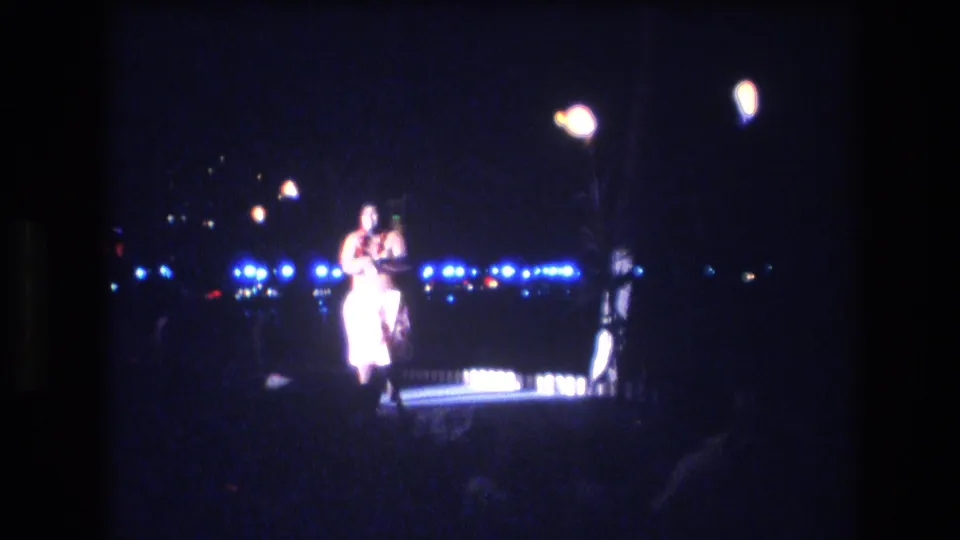 The performer remains centered under bright stage lamps, with the surrounding audience area mostly dark and blue lights visible in the background.