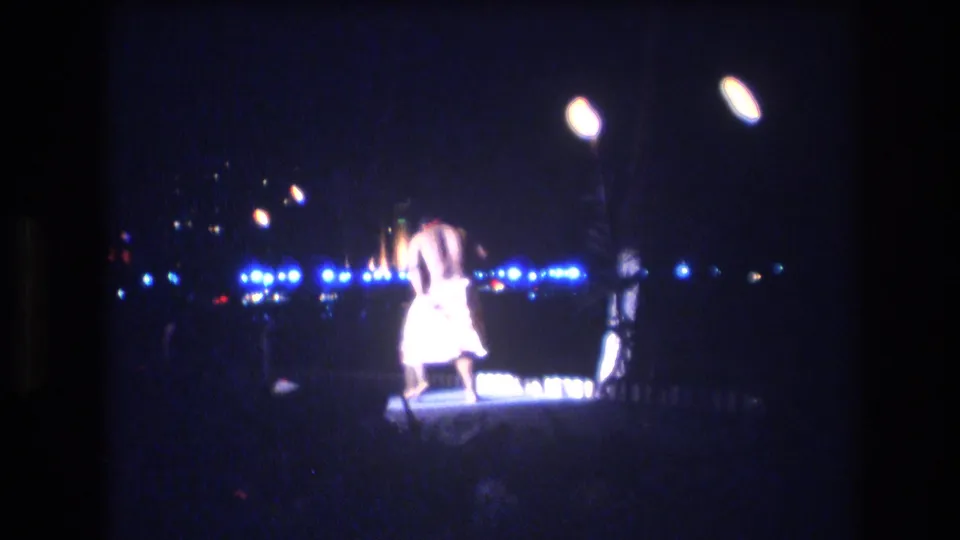 The male hula dancer is seen from behind or three-quarter back view on the dimly lit stage, with blue lights stretching across the nighttime background.