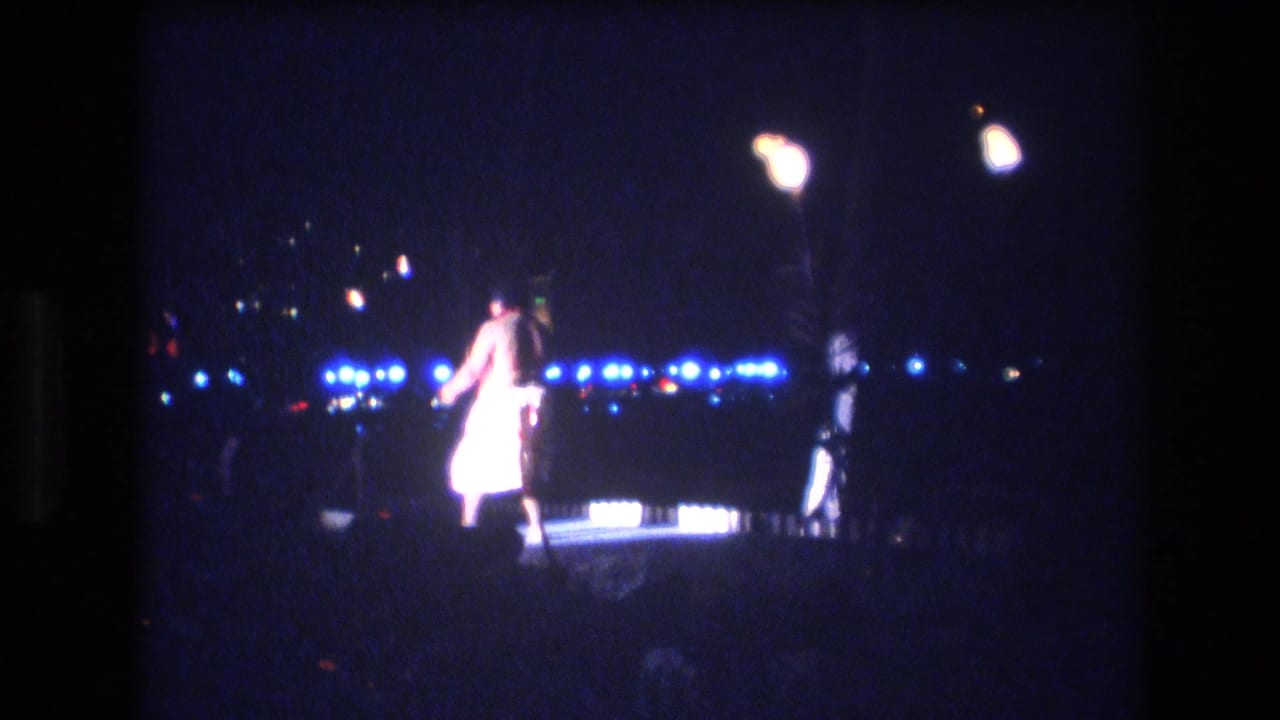 A shirtless adult dancer in light shorts stands on a dimly lit Honolulu stage at night in 1977, with blue lights and palm shapes visible in the background.