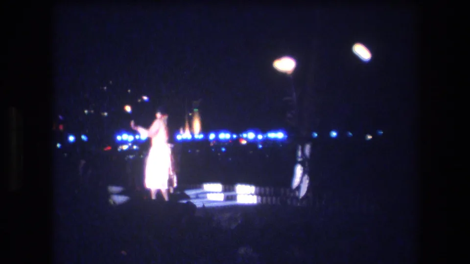 The performer gestures while standing under bright white lamps and blue stage lights, with the Honolulu night setting mostly dark around him.