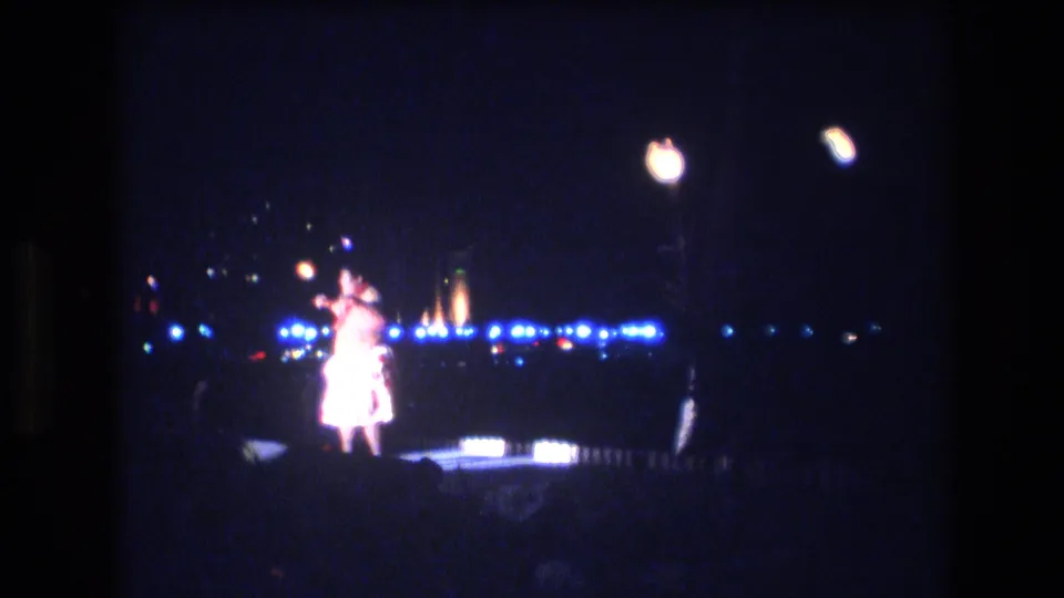 The dancer appears overexposed as he moves on the dark 1977 Honolulu stage, with blue lights, palm silhouettes, and distant points of light behind him.
