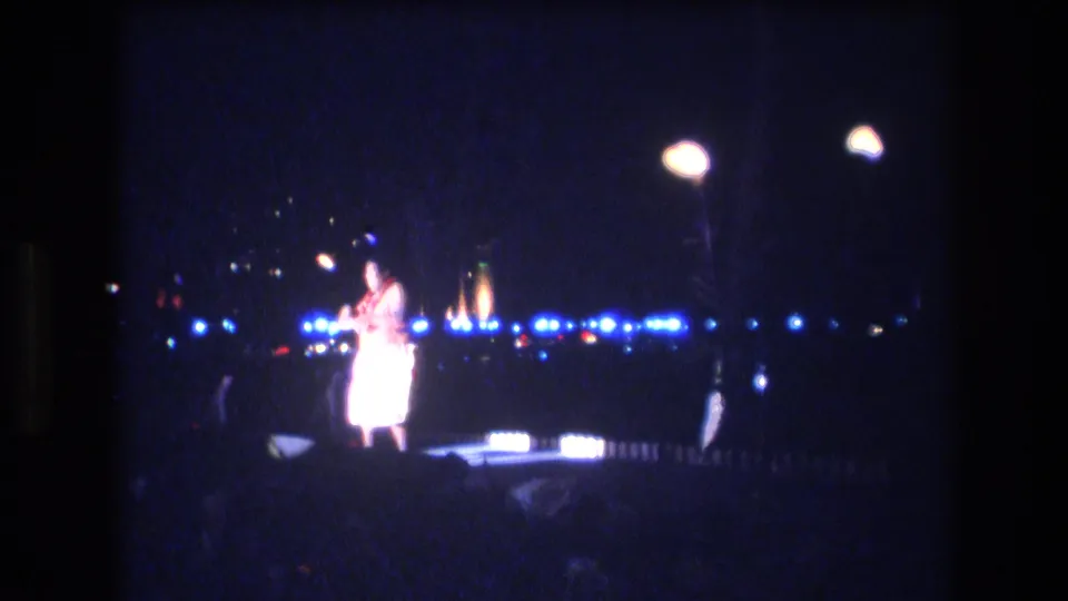 A shirtless adult dancer remains near center frame on a dark Honolulu stage, surrounded by blooming blue and white lights in grainy 8mm footage.