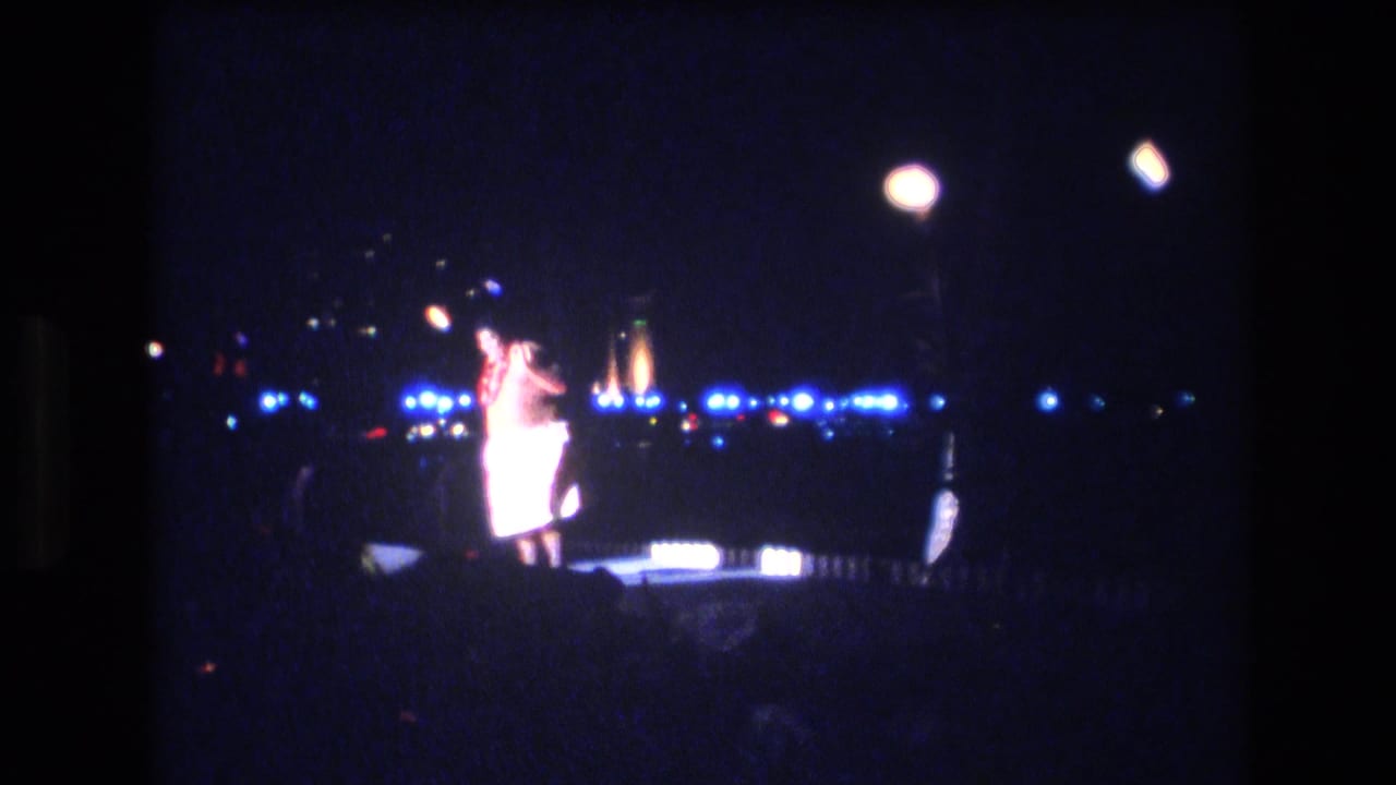 In 1977 Honolulu luau footage, a male performer in a light costume stands on a small night stage with blue lights, overhead lamps, and torch-like flames in the background.