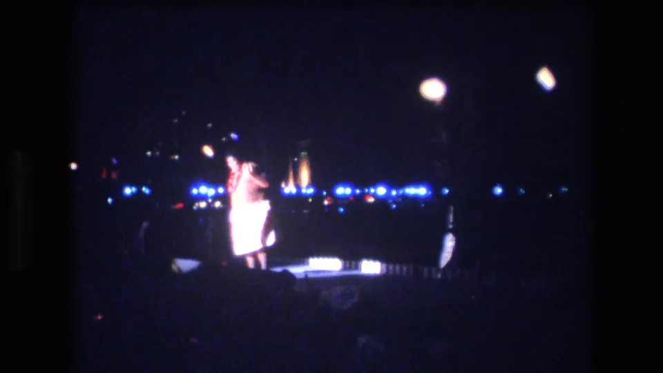 In 1977 Honolulu luau footage, a male performer in a light costume stands on a small night stage with blue lights, overhead lamps, and torch-like flames in the background.