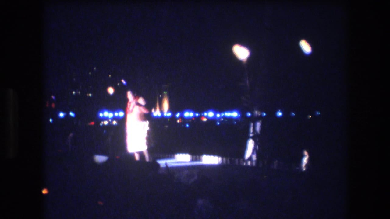 Performer gestures on a nighttime stage with bright lamps, blue light points, and torches in the distance.