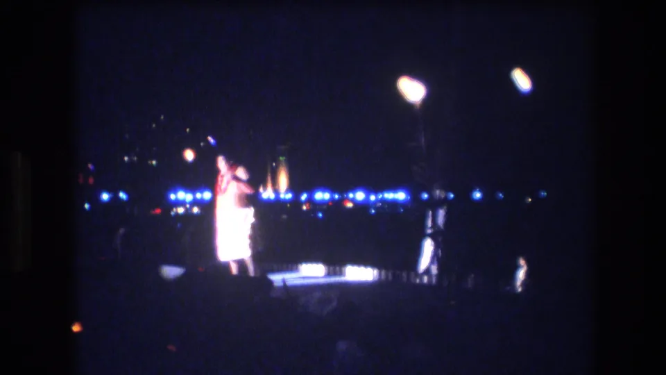 A male dancer appears mid-gesture on a nighttime Honolulu luau stage, framed by bright overhead lamps, blue stage lights, and distant torch-like flames.