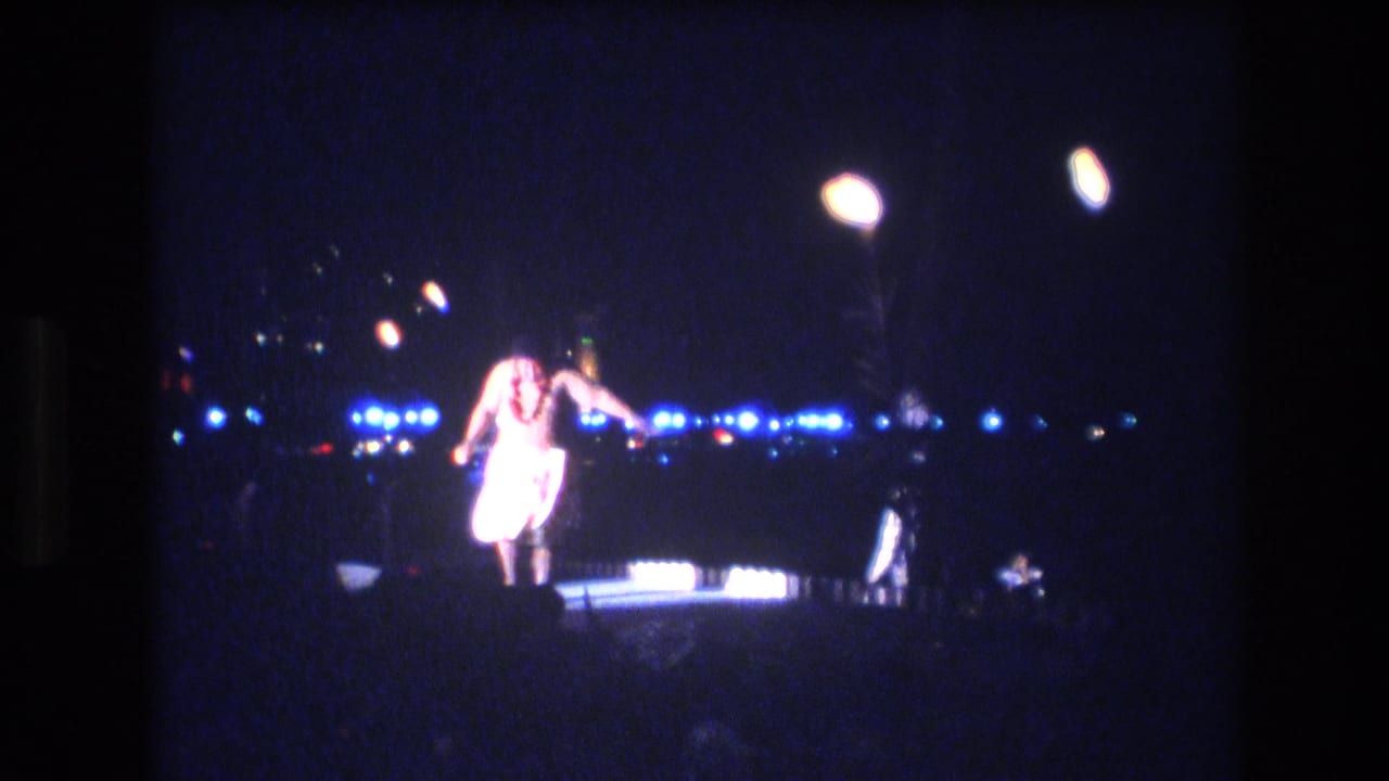 The 8mm frame shows the luau performer bending or stepping forward on the lit stage, with blue lights and torch-like flames visible across the night backdrop.