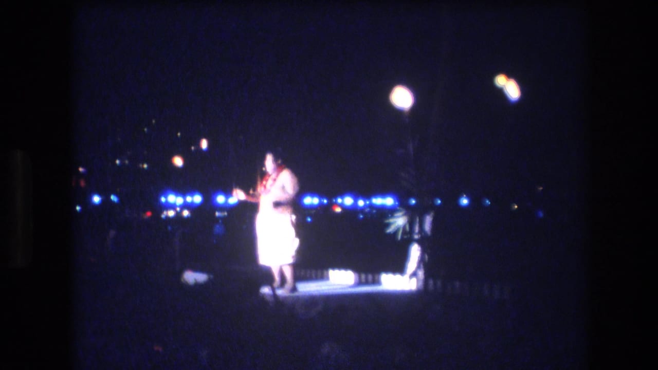 A male luau performer stands near center stage in the 1977 Honolulu night scene, illuminated by overhead lamps with blue lights and torch-like flames behind him.