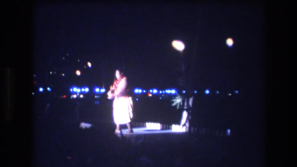 The performer remains centered on the lit platform, with the dark Honolulu night setting and rows of blue distant lights visible behind him.
