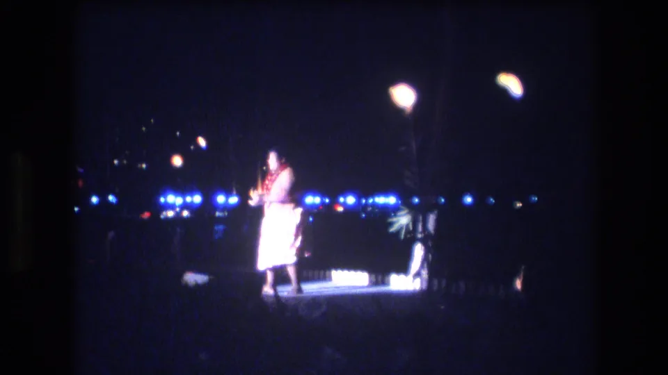 A handheld 8mm view shows the luau dancer on the lit platform, with pronounced grain, soft focus, and night lights beyond the stage.
