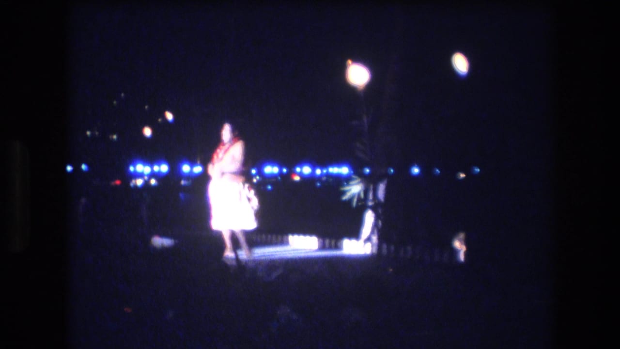 In 1977 Honolulu, Oahu home-movie footage, a dancer in a pale costume and red lei appears under bright lamps with blue lights across the dark background.
