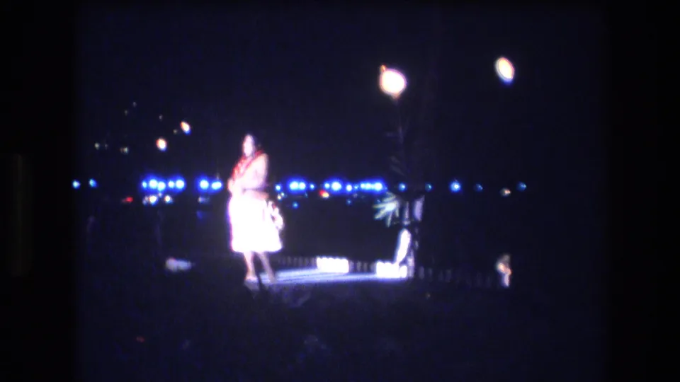 In 1977 Honolulu, Oahu home-movie footage, a dancer in a pale costume and red lei appears under bright lamps with blue lights across the dark background.