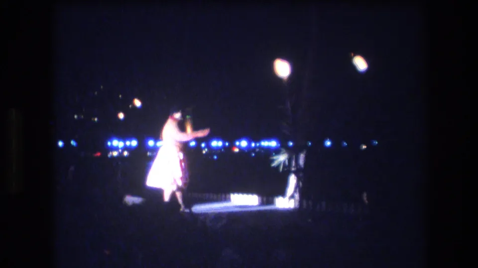 The Honolulu luau dancer is seen mid-gesture in a light-colored outfit, filmed from a distant audience position against blue lights and night sky.