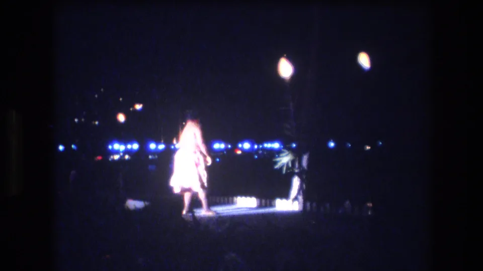 A light-clad dancer continues the luau performance on a small illuminated stage, with blue lights and faint palm-like shapes in the nighttime background.