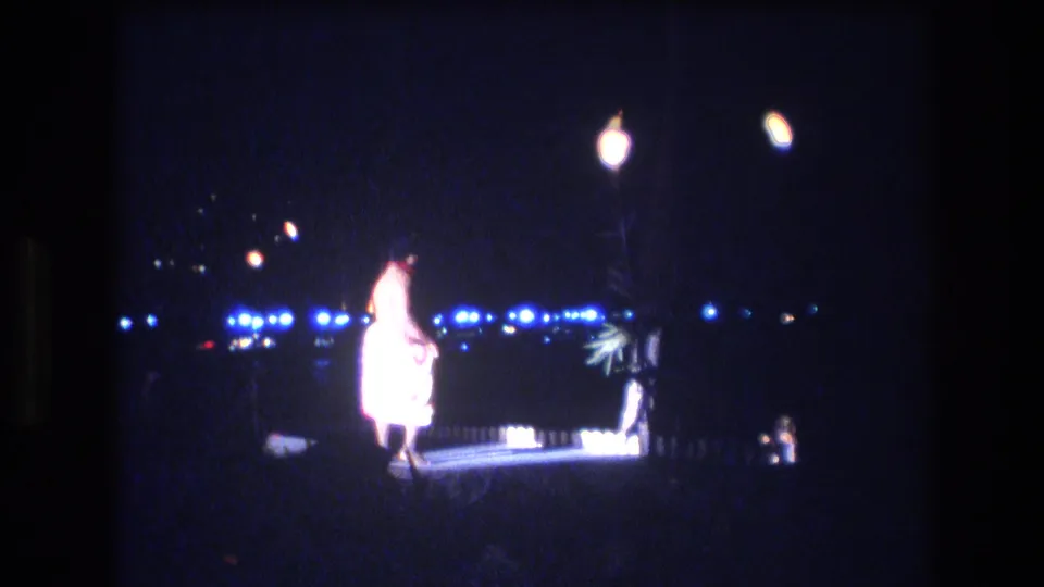 The final frame holds on the 1977 Honolulu luau performer near center stage, surrounded by bright lamp bloom, blue background lights, and dark outdoor space.