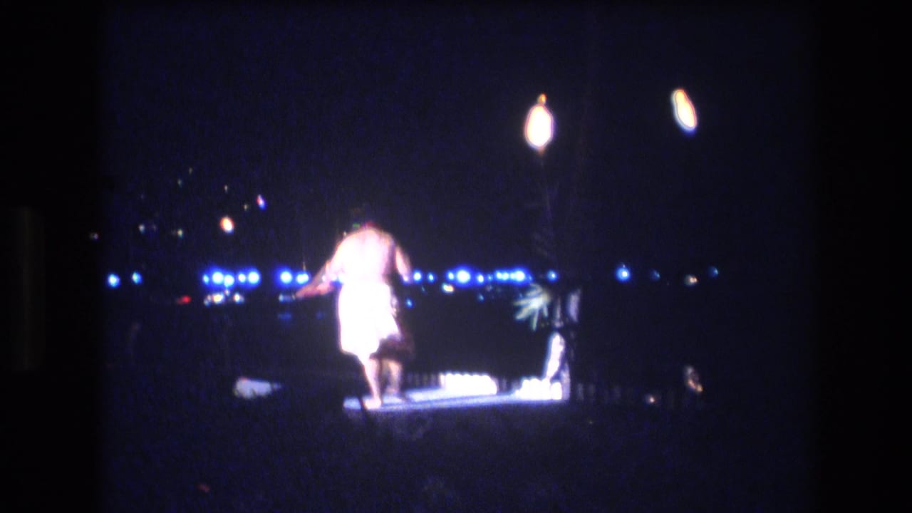 Dancer in a light garment on a small outdoor stage at night with bright distant lights.