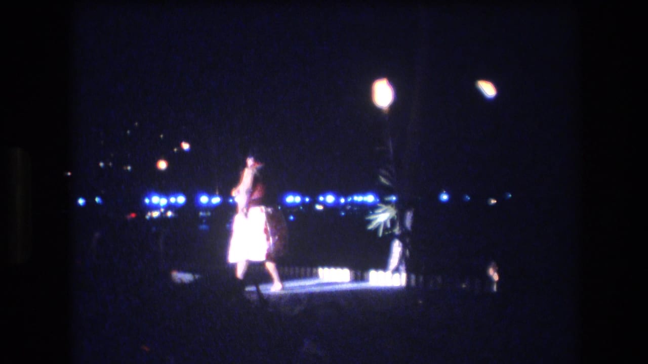 Performer raises an arm on a dark outdoor stage with a bright lamp and distant lights behind.