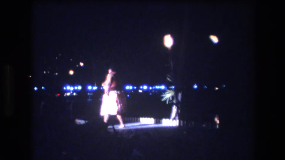 1977 Honolulu frame of the nighttime performer turning with one arm raised, silhouetted by stage lighting against a dark backdrop with city or waterfront lights in the distance.