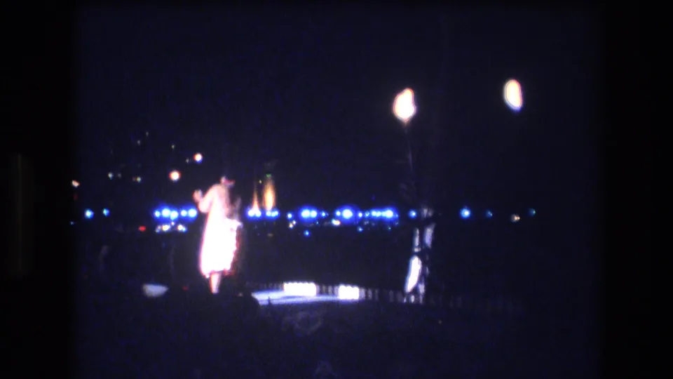1977 Honolulu home-movie frame showing the dancer from behind or in profile on the small lit stage, with bright lamps above and a line of distant blue-white night lights behind the performance area.