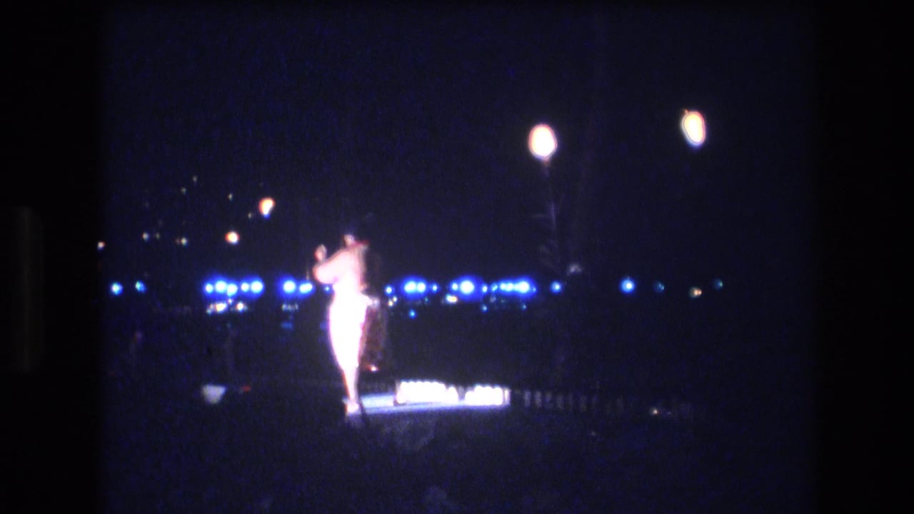 A lone dancer in a light-colored costume performs at night in 1977 Honolulu footage, framed from a distant handheld audience viewpoint with blue lights behind her.
