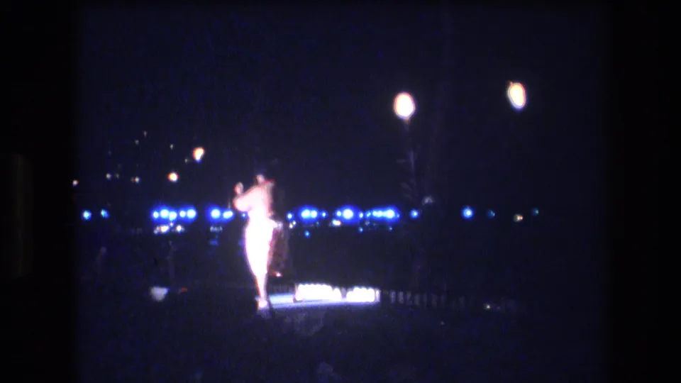 A lone dancer in a light-colored costume performs at night in 1977 Honolulu footage, framed from a distant handheld audience viewpoint with blue lights behind her.