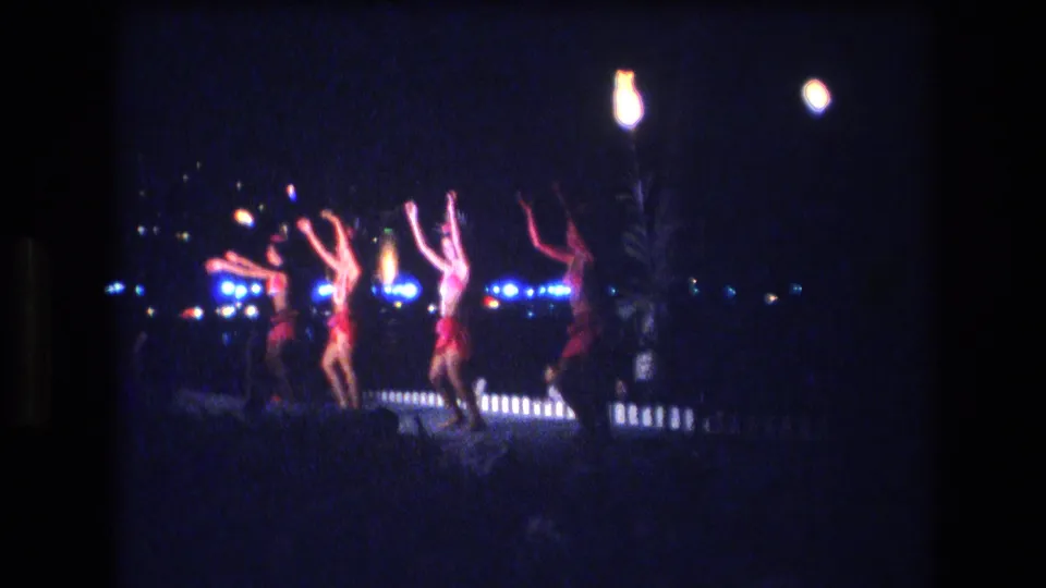 A group of hula dancers in red and pink-toned costumes perform in unison under bright lamps and blue background lights in nighttime Honolulu home-movie footage.