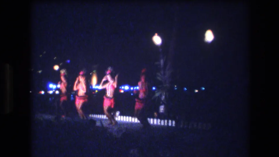 Several performers in red and pink costumes continue a synchronized nighttime dance routine, with torch-like lamps and blue lights visible behind them in 1977 Honolulu footage.
