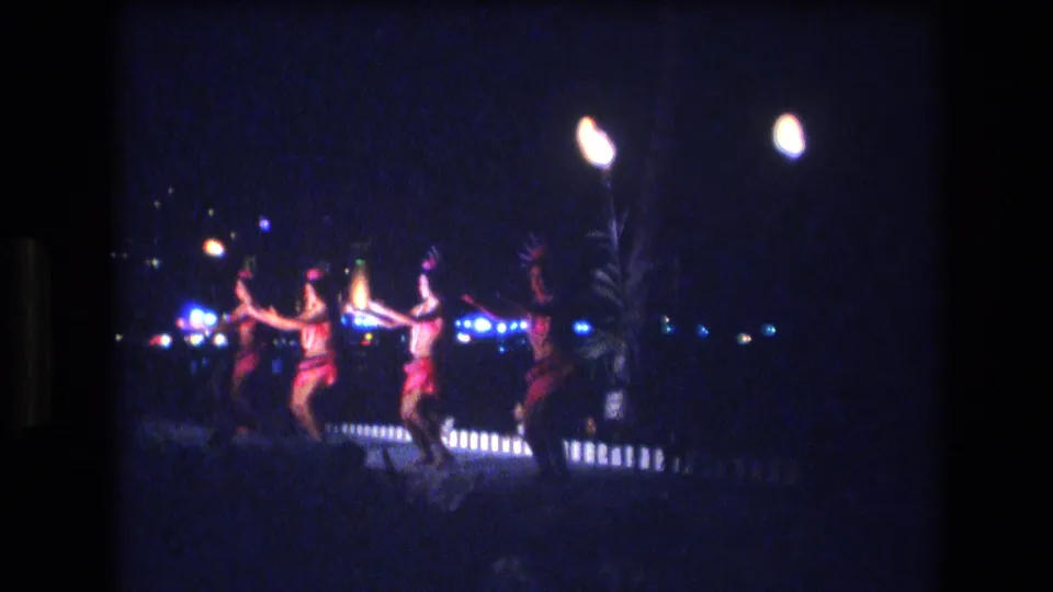 Dancers in red-toned costumes extend their arms during a nighttime hula-style performance, seen from a distant handheld 8mm perspective in Honolulu.