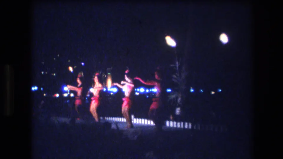 Women in red and dark costumes perform in a distant nighttime stage view, with bright lamps and small blue lights visible behind them in Honolulu footage dated 1977.
