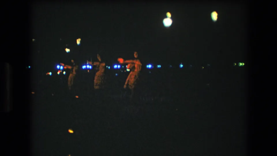 The dancers are dimly visible in a low-light 8mm frame, with small blue and green lights and a few warm torch-like points against the night.