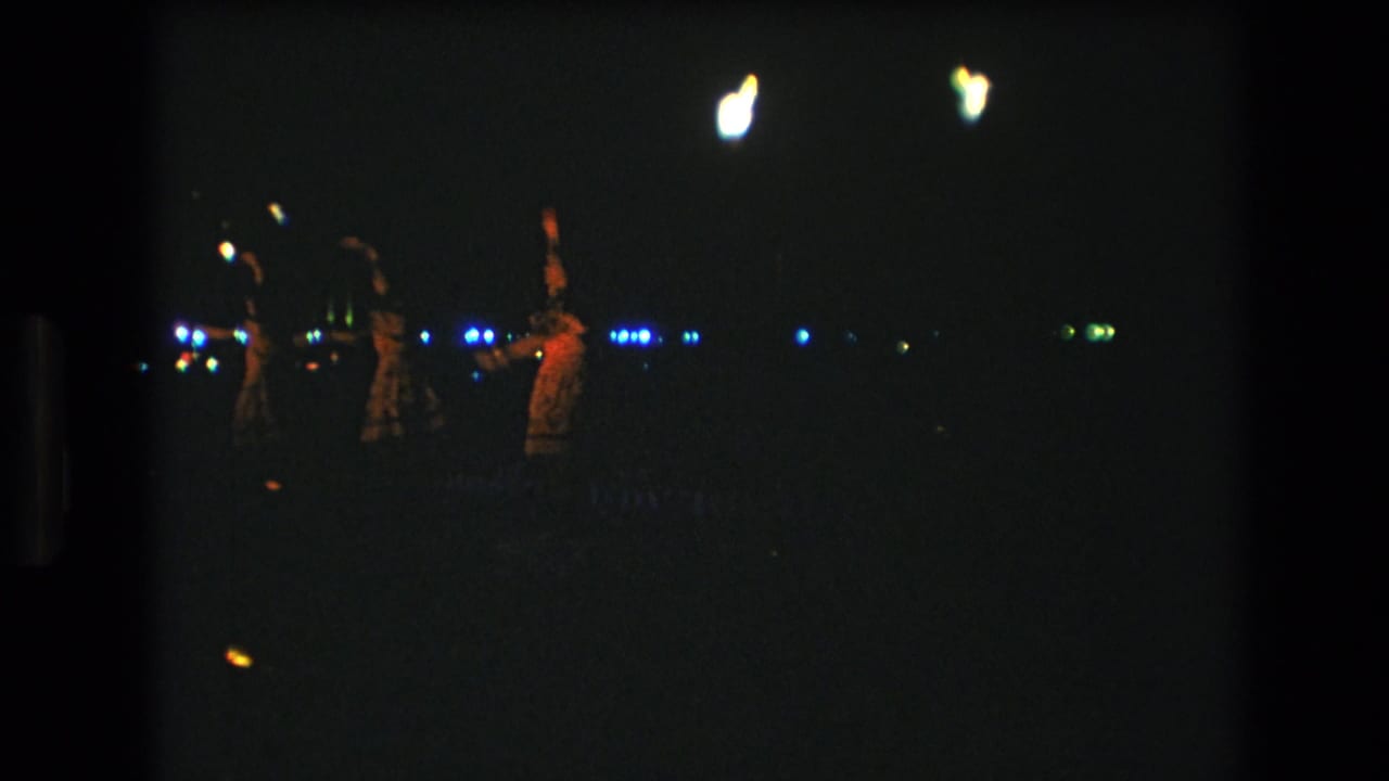 Distant nighttime dancers lit by small flames and background lights.