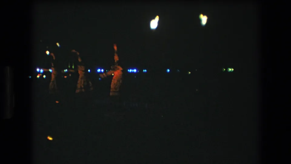 1977 Honolulu home-movie frame showing distant dancers at a nighttime luau-style gathering, with warm torchlight and blue background lights in a dark scene.
