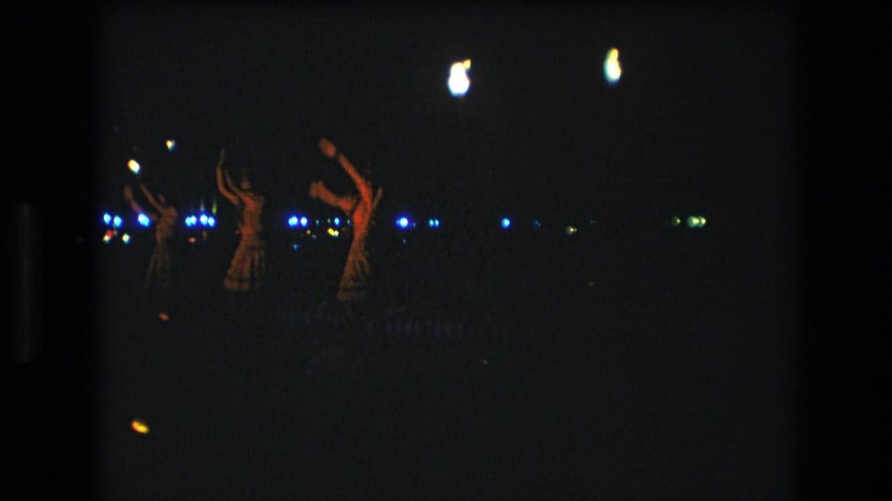 1977 Honolulu frame with several distant performers raising their arms during a nighttime luau-style dance, seen through grainy 8mm film exposure.